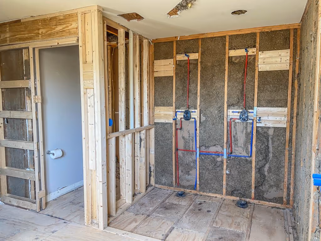 Bathroom under construction with exposed wooden framing, plumbing pipes in red and blue, and unfinished walls.