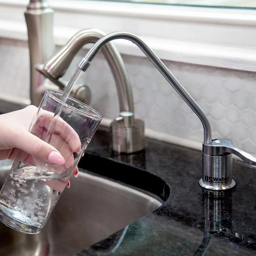 Hand holding a glass under a stainless steel water filter faucet filling it with clear water over a kitchen sink.