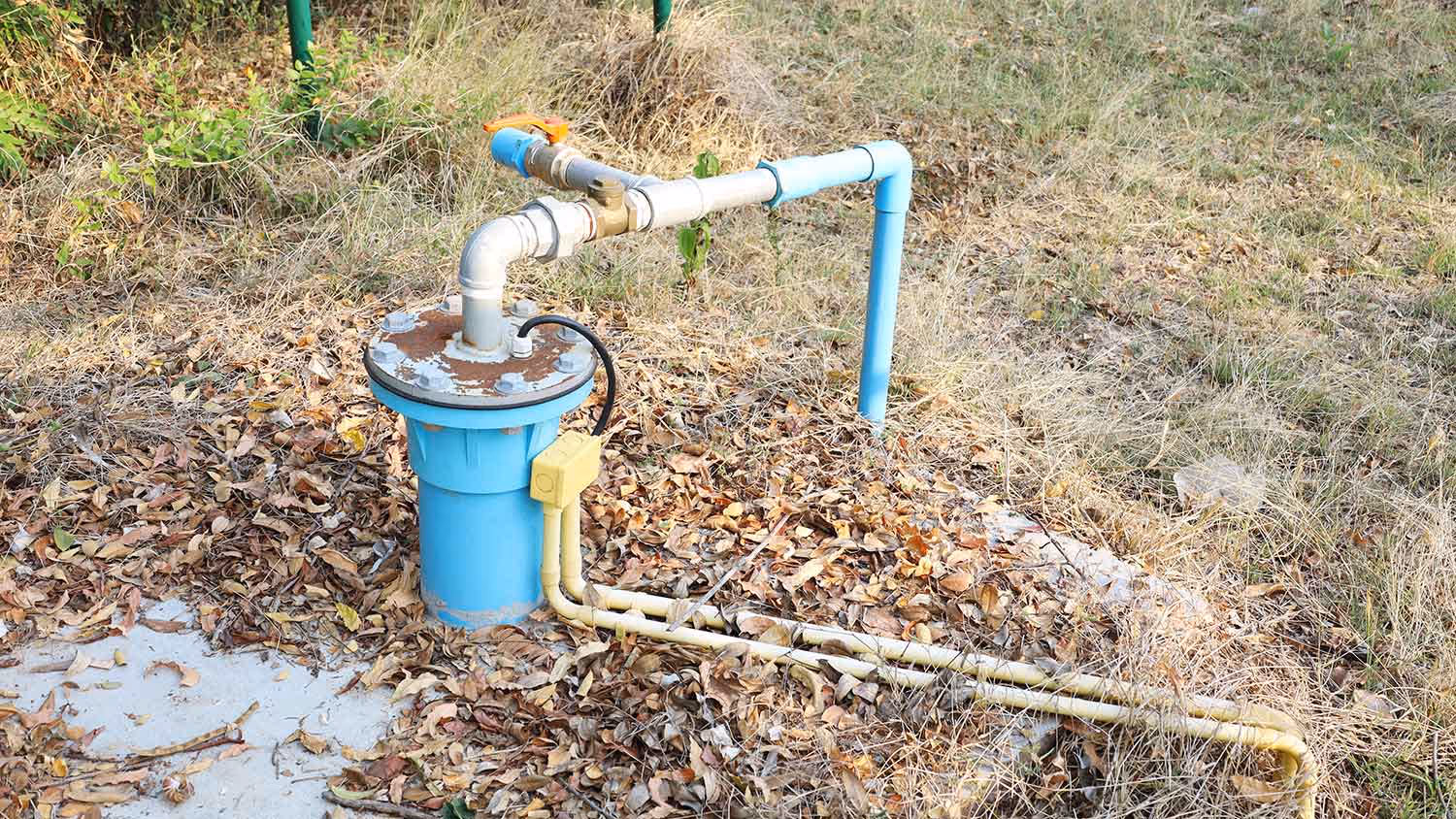 Blue submersible well pump with connected pipes installed in dry grassy area covered with fallen leaves.
