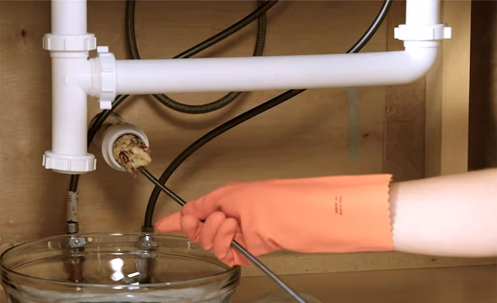 Person wearing an orange glove using a drain snake to clean debris from a clogged drain pipe under a sink.