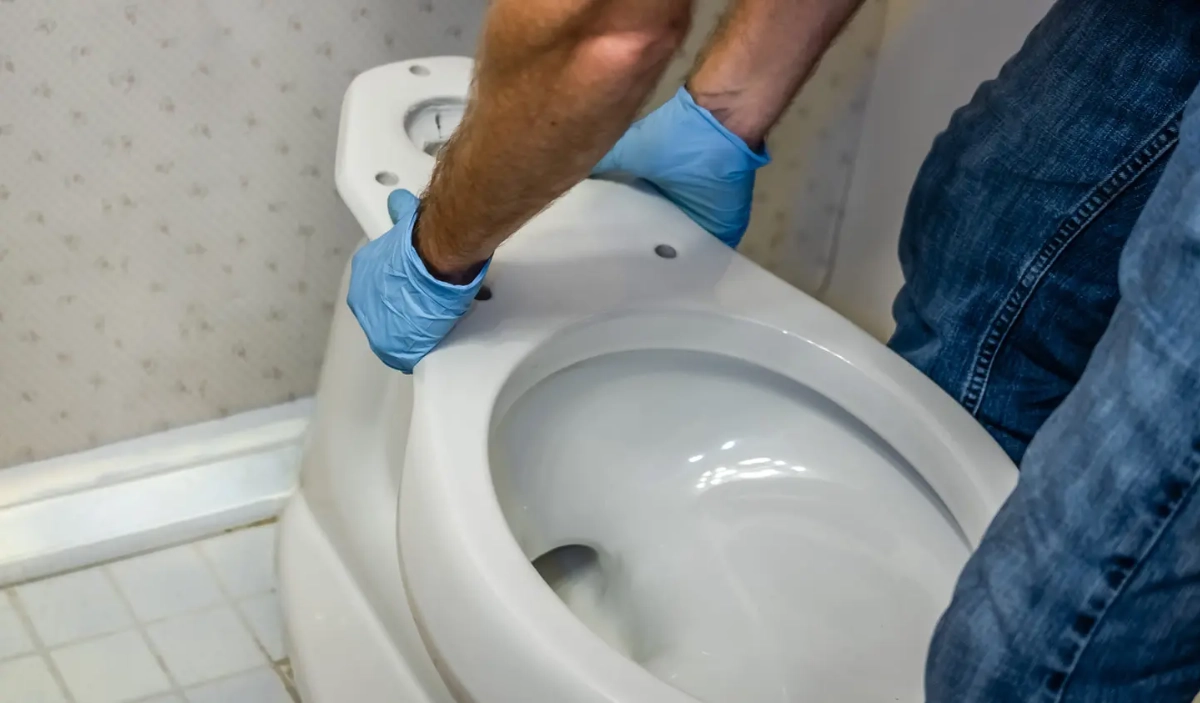 Person wearing blue gloves replacing or installing a white toilet bowl indoors  in Worthington, Ohio.