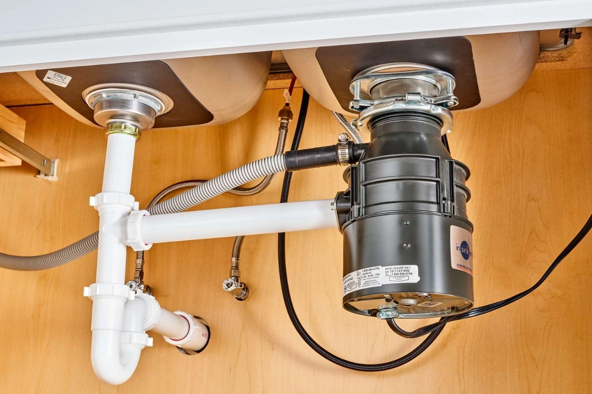 Under-sink view showing dual sink drains connected to a garbage disposal unit with white PVC piping and water valves on a wooden cabinet wall in Hilliard, Ohio.