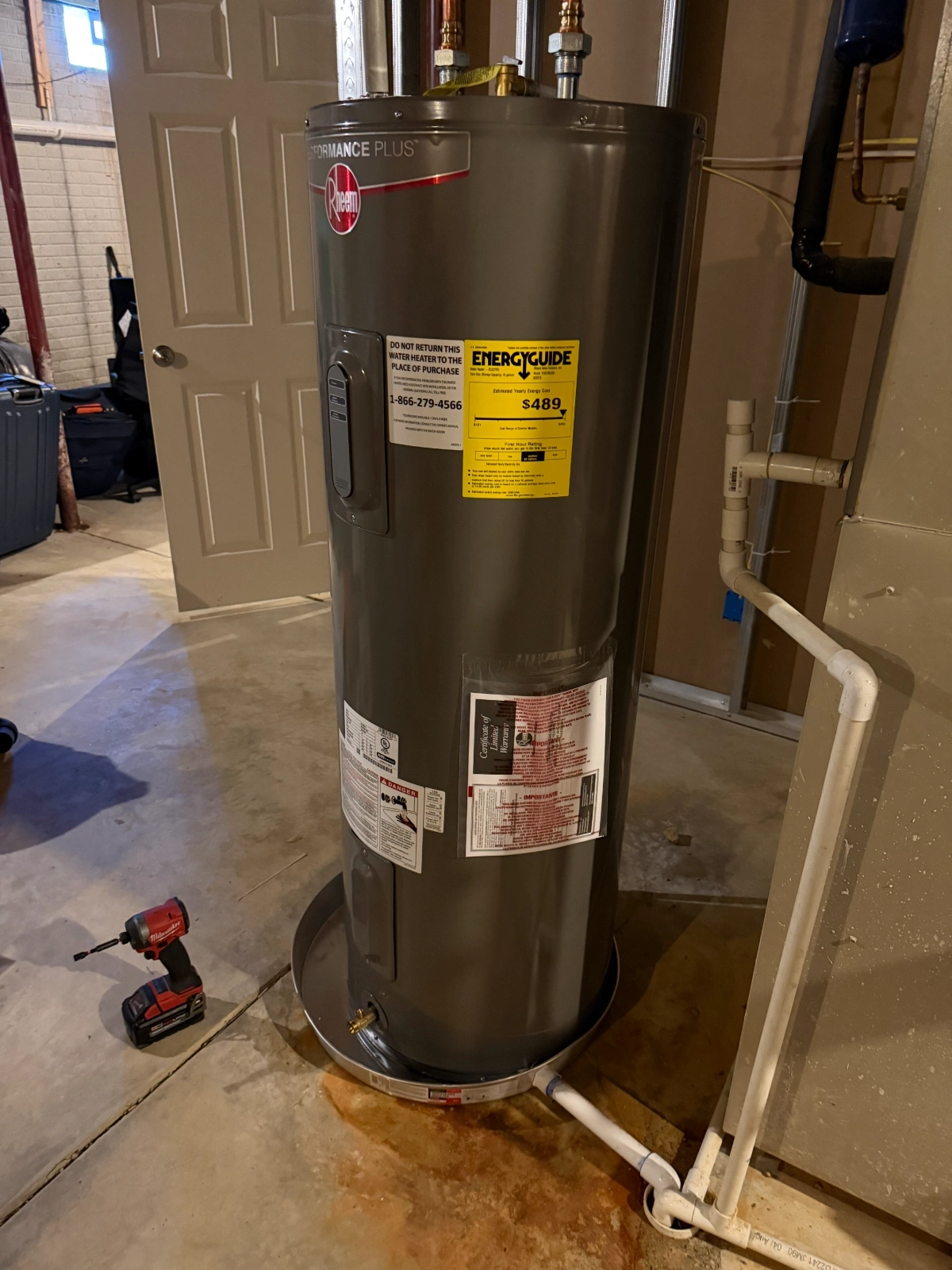 Tall gray Rheem Performance Plus water heater with energy guide label, connected to white plumbing pipes, in a basement with a concrete floor and a red cordless drill nearby in Hilliard, Ohio.