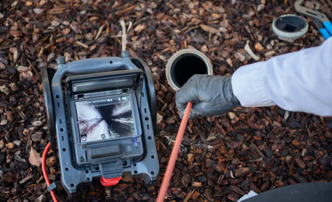Worker using a drain inspection camera displaying the inside of a pipe on its screen, with the camera positioned on mulch ground in Dublin, Ohio.