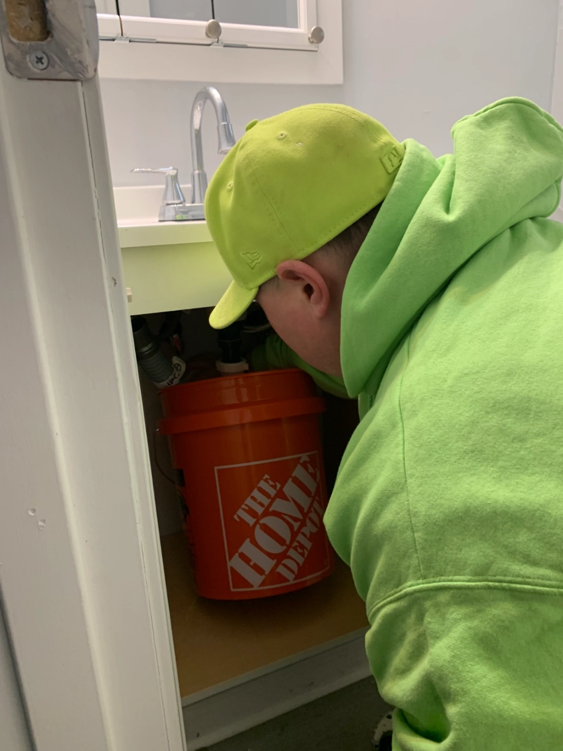 Person in neon green hoodie and hat working under a bathroom sink with a Home Depot bucket inside the cabinet in Dublin, Ohio.
