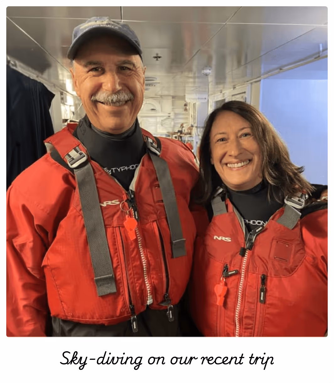Smiling man and woman wearing red life jackets and black wetsuits inside a boat or ship cabin.