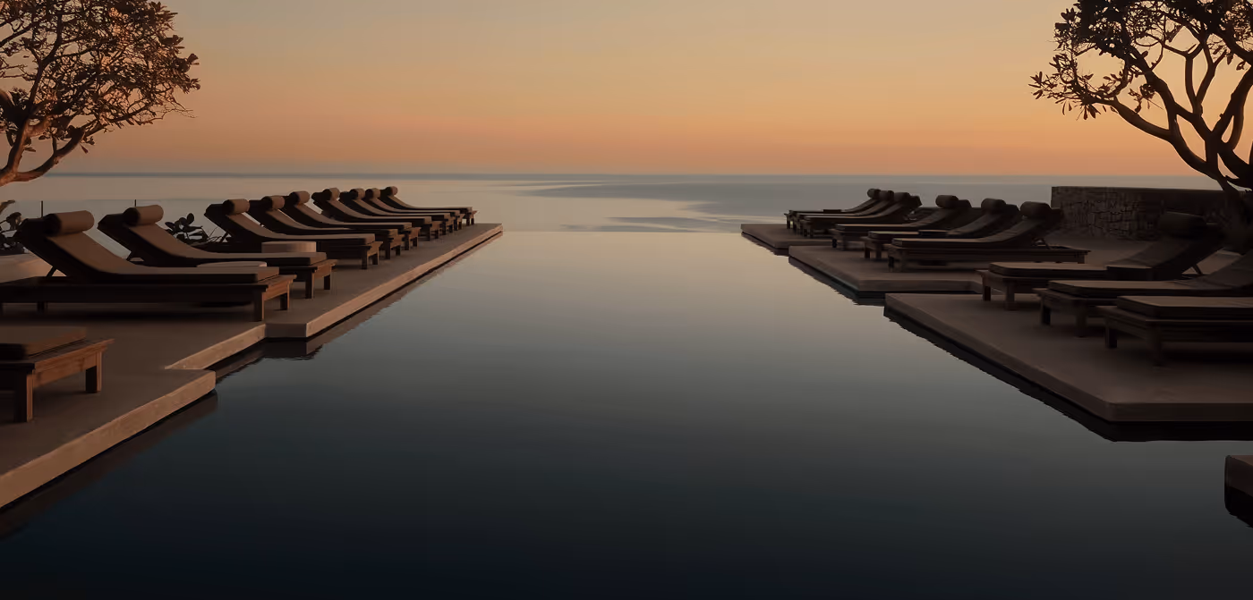 Infinity pool with lounge chairs on both sides overlooking calm ocean during sunset.