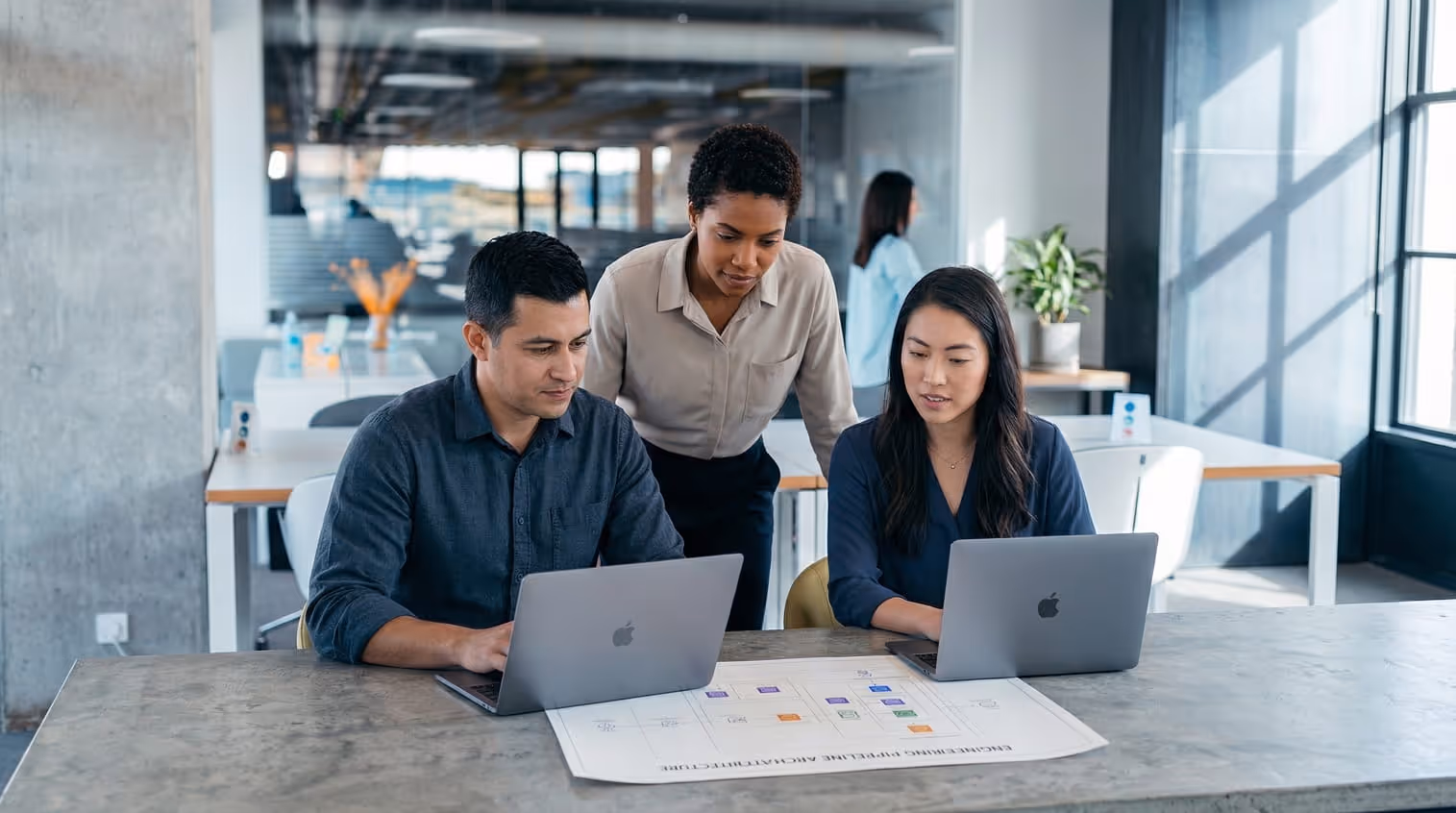 Three colleagues collaborating in an office at a table with laptops and a printed diagram.