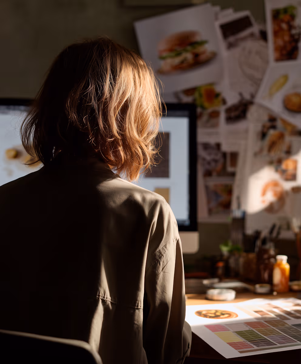 Person with shoulder-length hair sitting at a desk with design prints and a computer screen in a dimly lit creative workspace.