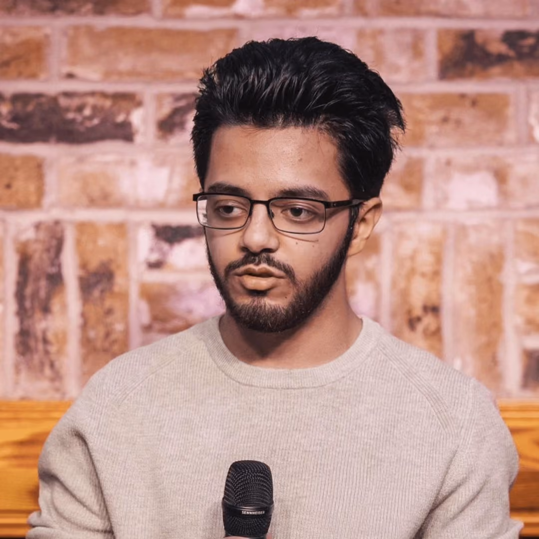 Young man with glasses holding a microphone against a brick wall background.