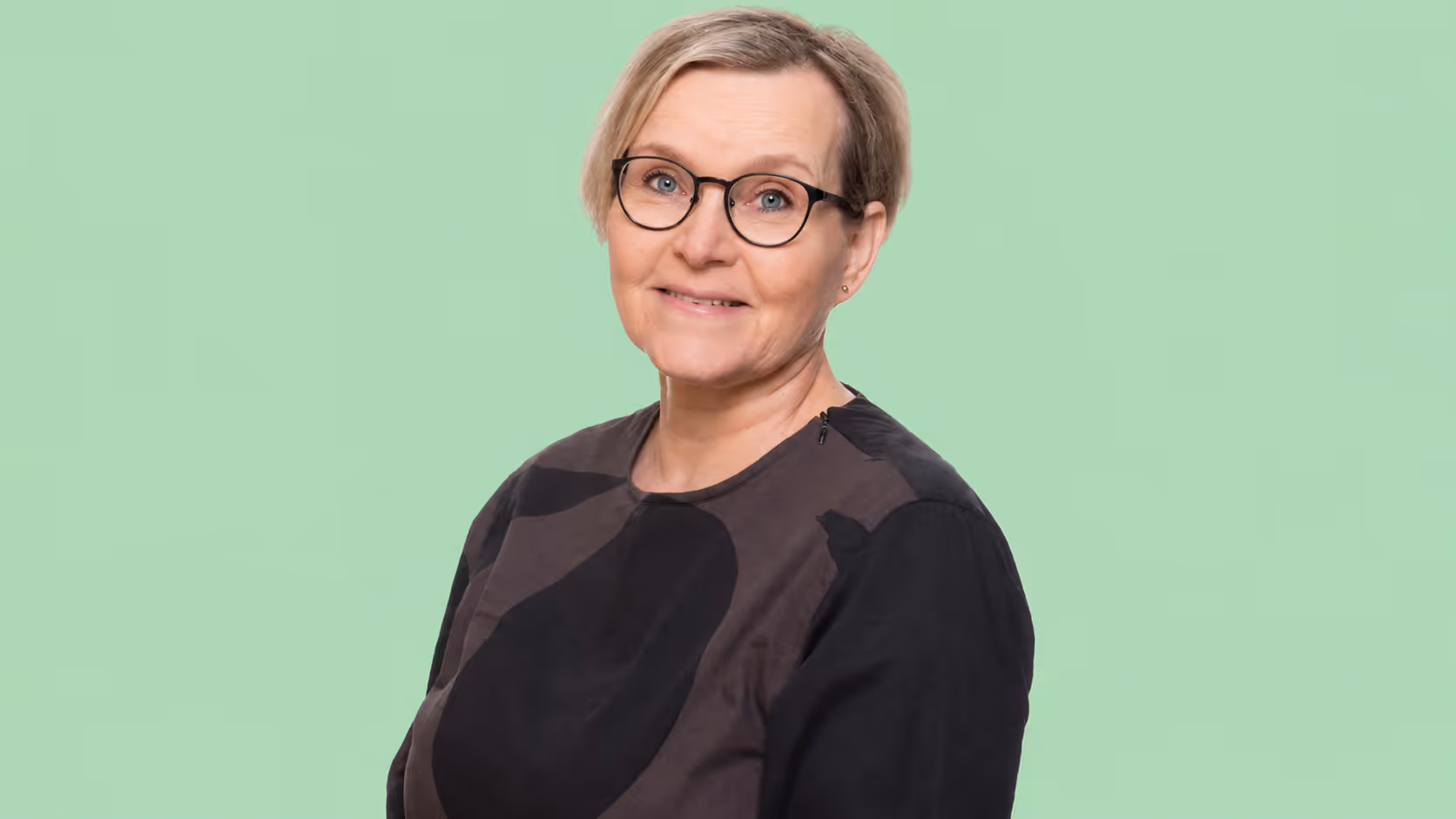Hymyilevä nainen, lyhyet vaaleat hiukset ja silmälasit vaaleanvihreää taustaa vasten.