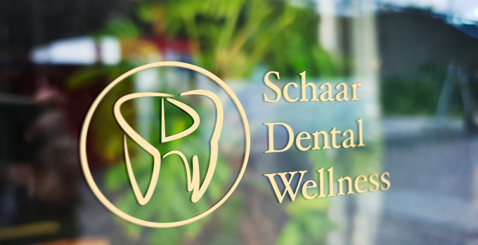 Gold tooth logo and the words ‘Schaar Dental Wellness’ displayed on a glass door with greenery softly visible in the background.