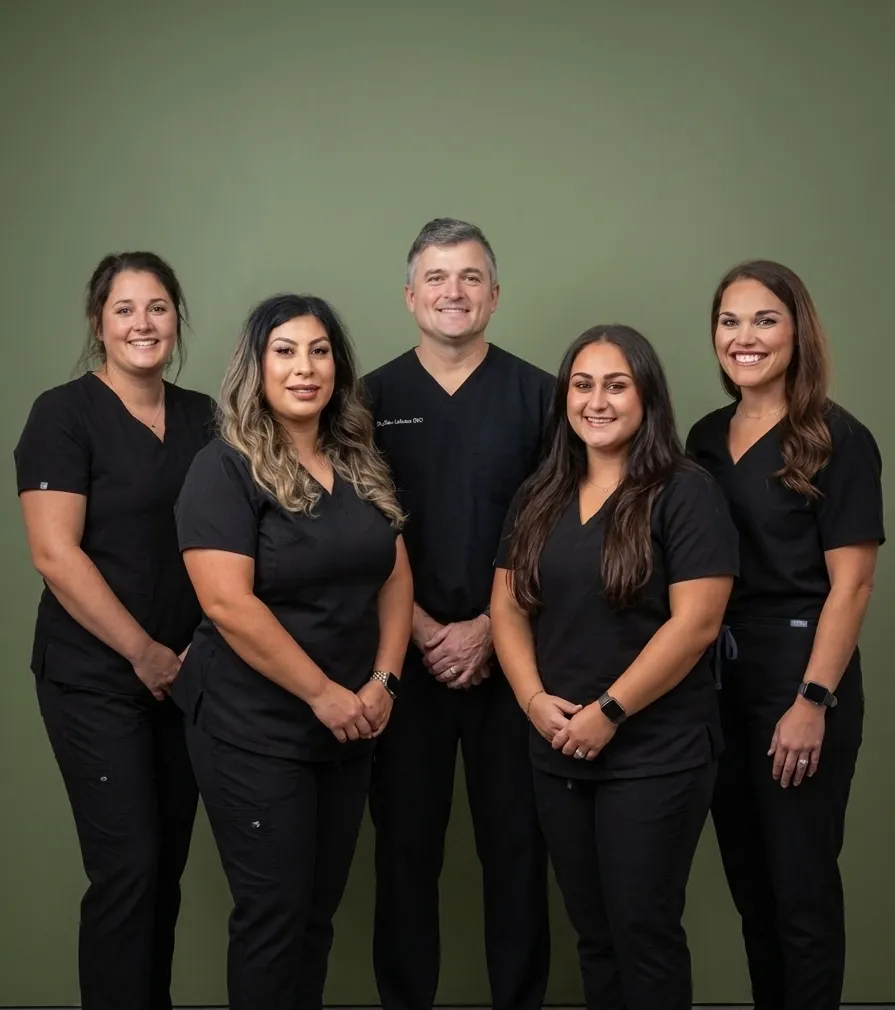 The Schaar Dental Wellness team of five in black scrubs smiling together against a green backdrop in Victoria, TX.
