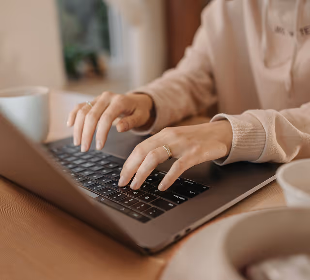 Person typing on a laptop keyboard at a wooden table with a coffee cup nearby.