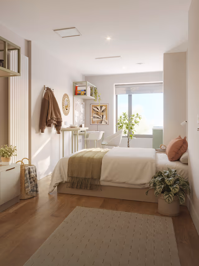Bright bedroom with wooden floor, a bed with white and beige bedding and pink pillows, plants, and a window with natural light.