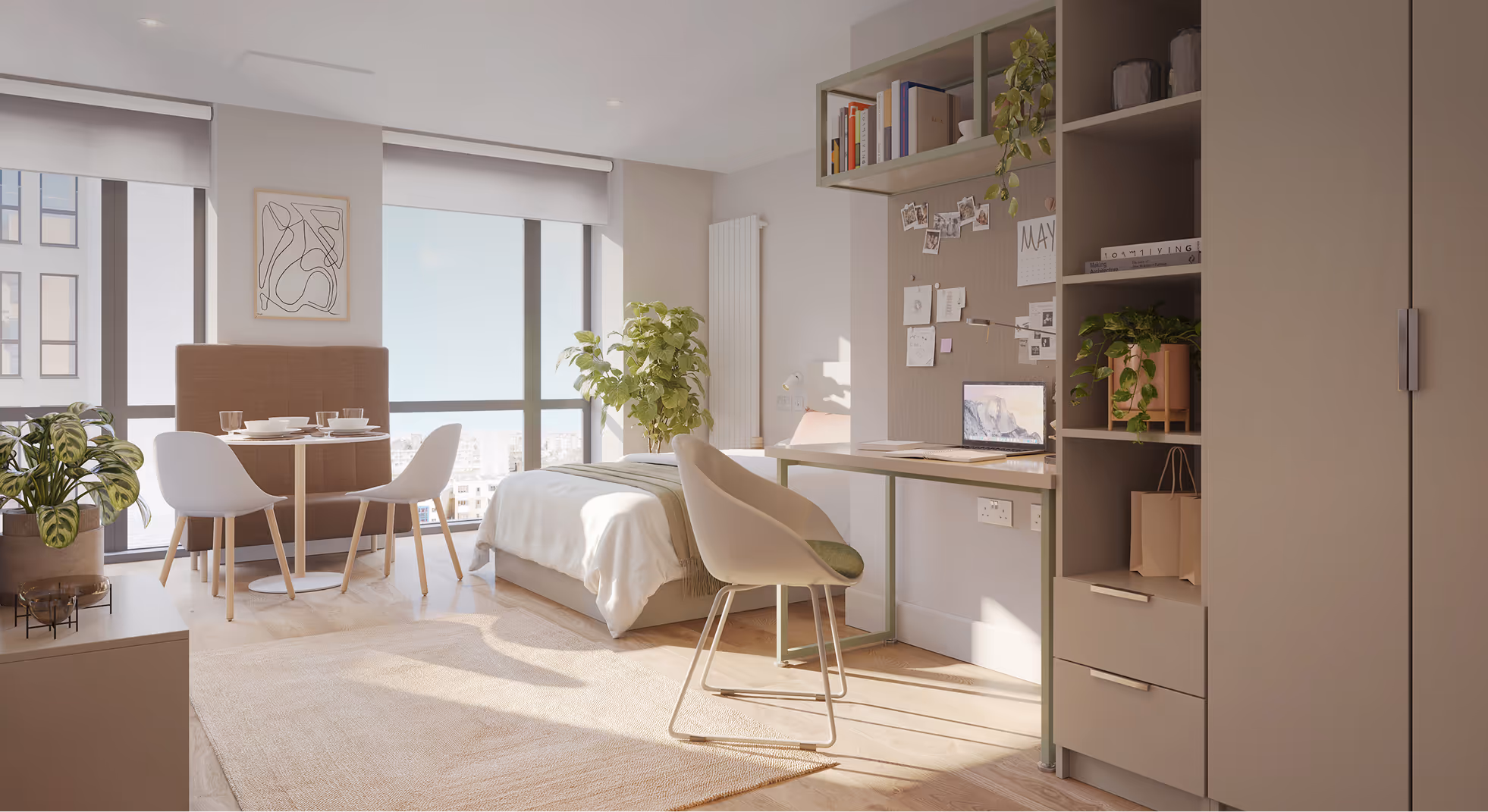 Bright modern studio apartment with large windows, a bed, dining table with two chairs, desk with a chair and laptop, and several green plants.