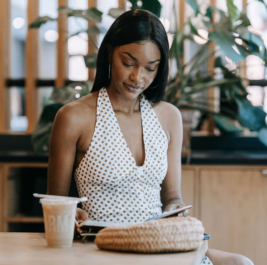 Woman in a white and brown polka dot halter dress reading a book at a wooden table with a drink and woven bag.