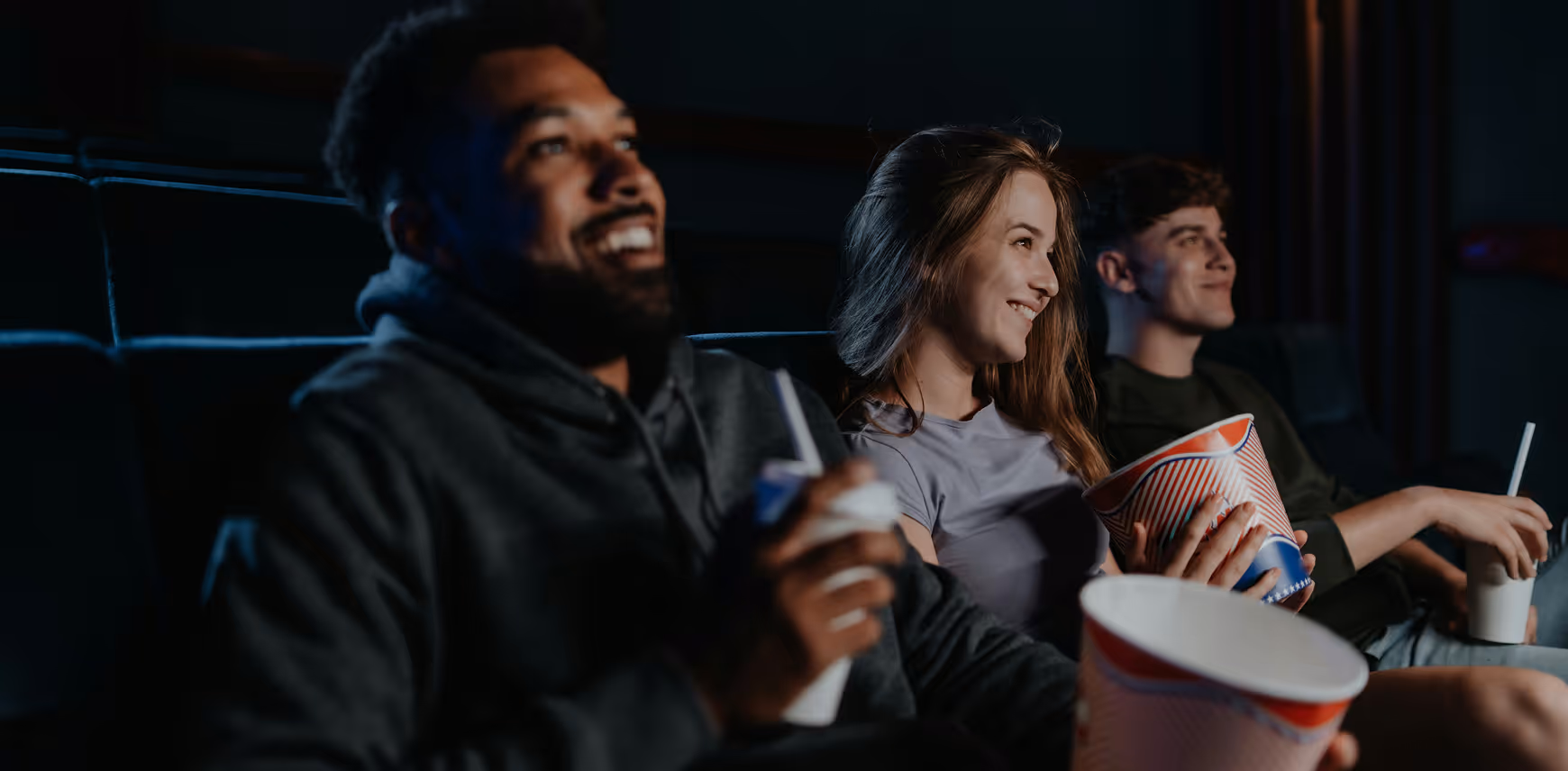 Three young adults smiling and enjoying a movie in a dark theater, holding popcorn buckets and drinks.