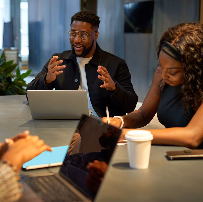 A man speaking and gesturing with hands during a meeting while a woman takes notes, with laptops and a coffee cup on the table.