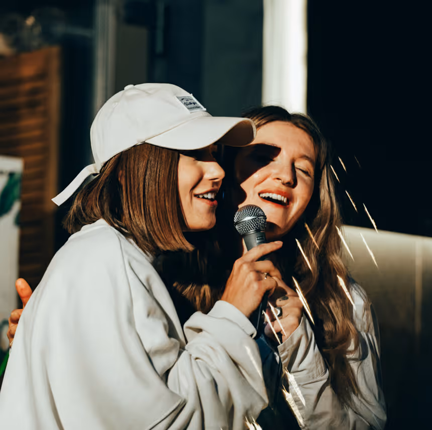 Two young women singing together into a microphone in a karaoke setting, one wearing a white cap and sweatshirt.