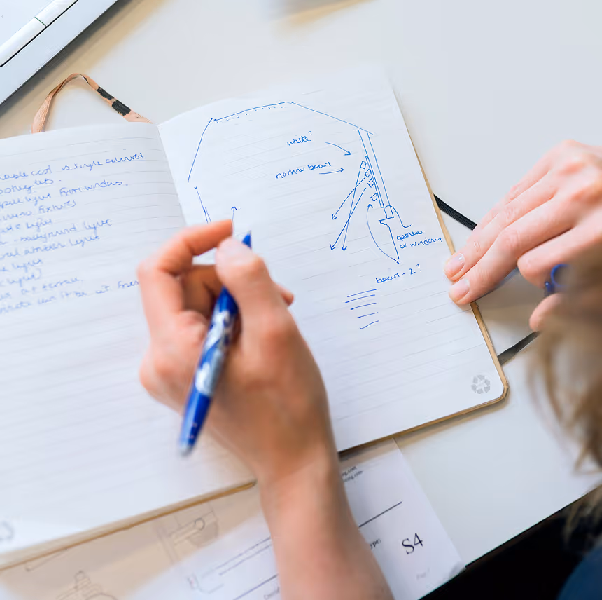 Person sketching a blueprint with handwritten notes in blue ink on a lined notebook.