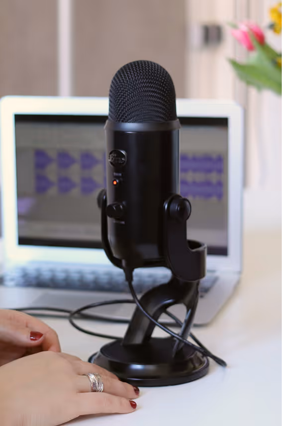 Black USB microphone on a desk in front of a laptop with audio editing software on screen.