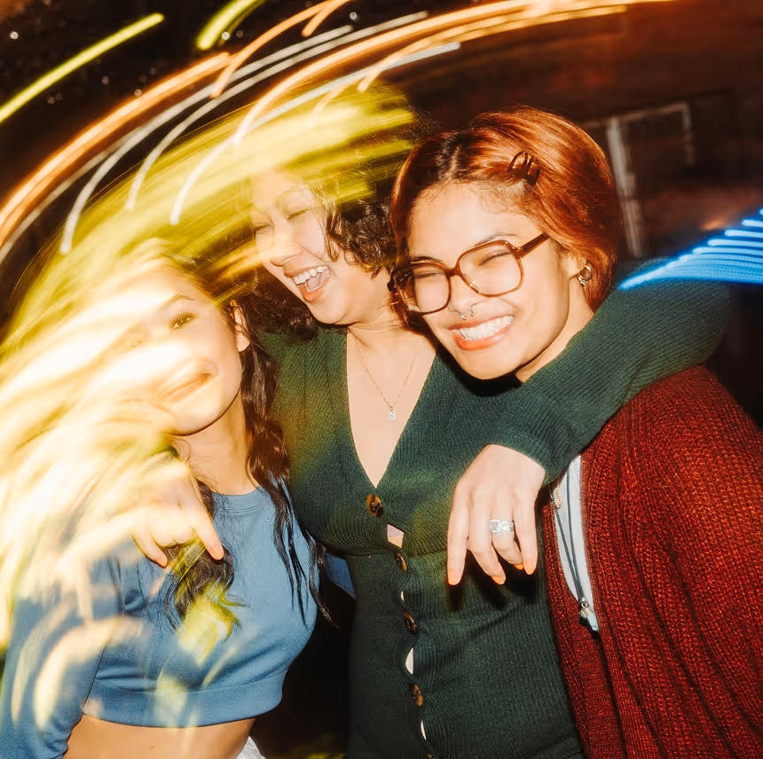 Three women smiling and embracing each other, with yellow and blue light streaks surrounding them.