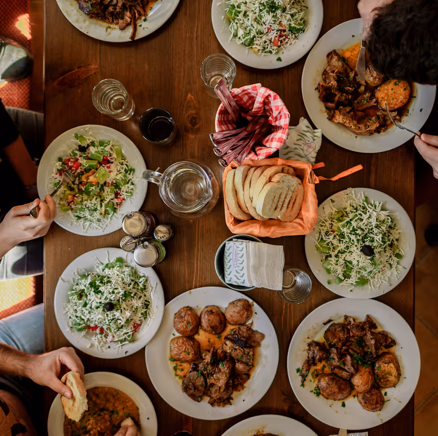 Overhead view of a wooden table set with plates of salad, cooked meat and potatoes, a basket of sliced bread, glasses of water, and people eating.