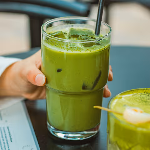 Hand holding a glass of iced matcha latte with a straw on a table, next to another matcha drink with a skewer of mochi balls.