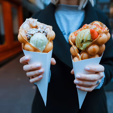 Person holding two bubble waffles filled with green ice cream, topped with strawberries and nuts, in white paper cones.