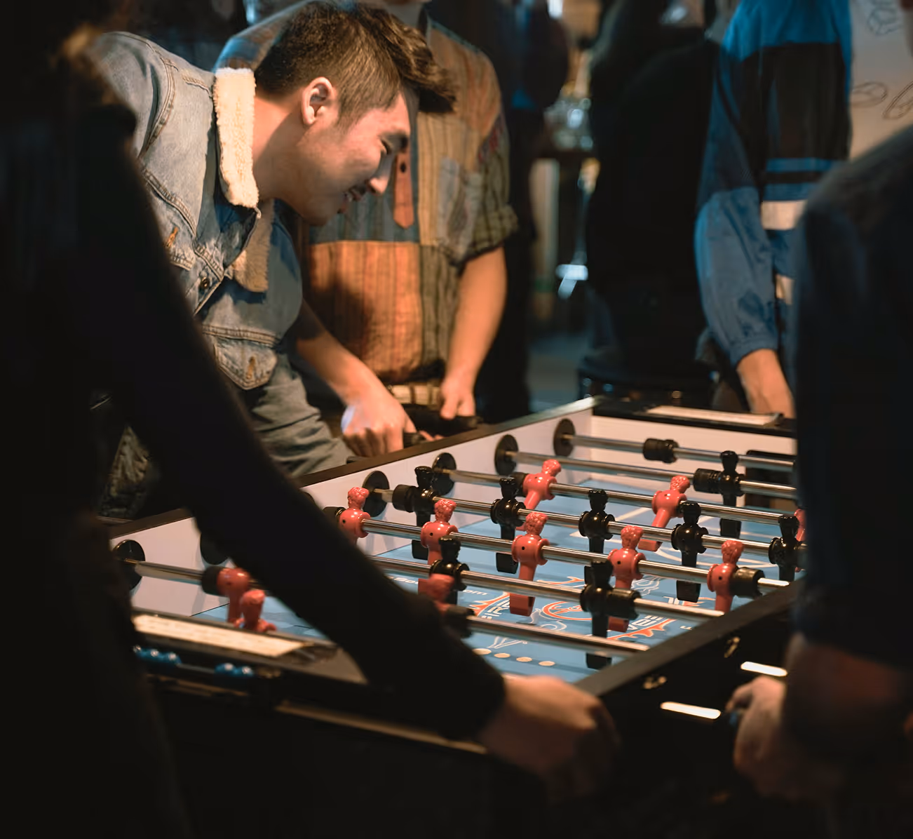 Young people playing foosball indoors, focusing on the foosball table and players controlling rods with black and red figures.