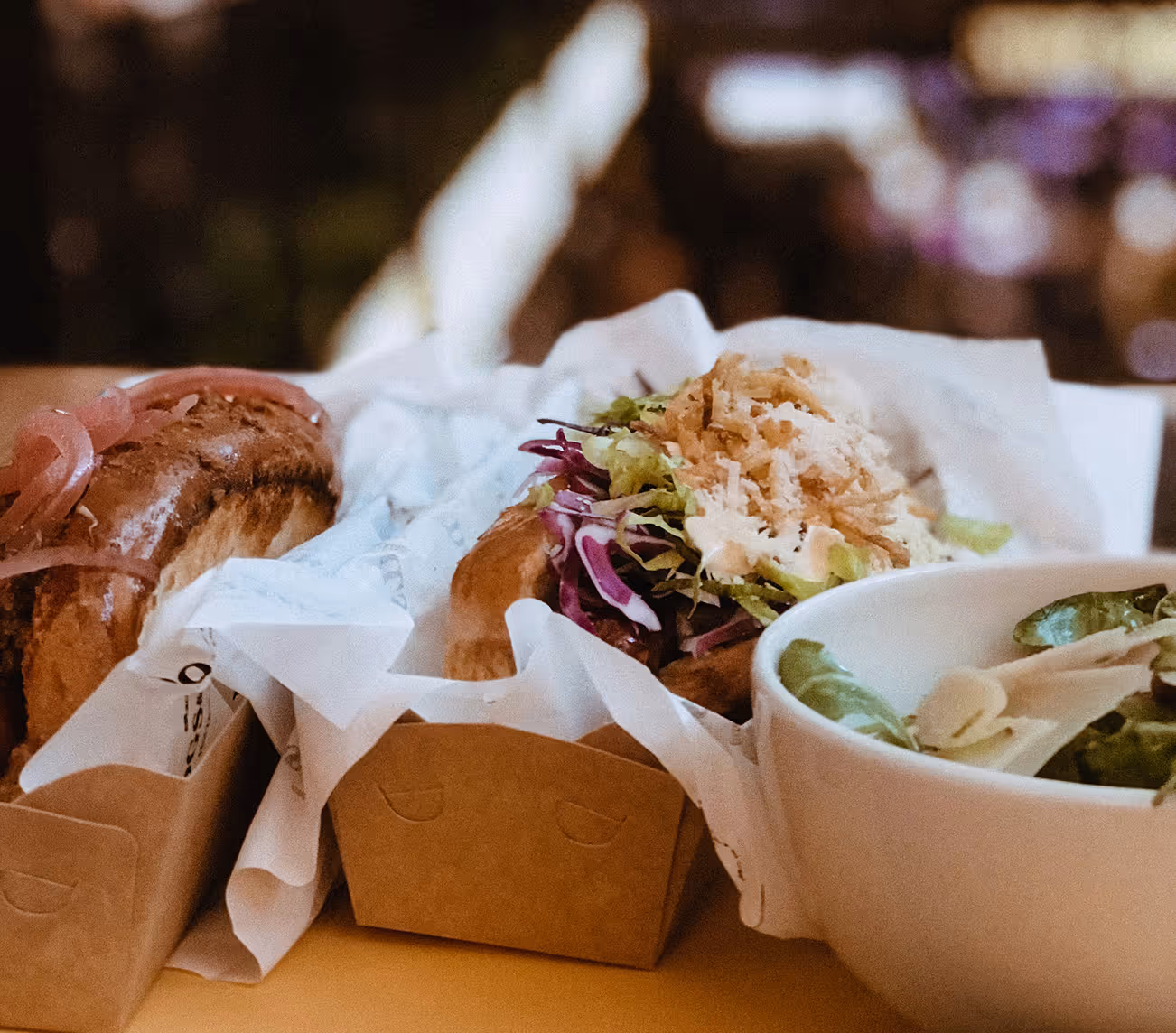 Two sandwiches with various toppings in brown paper trays next to a white bowl of salad with greens and sliced almonds.