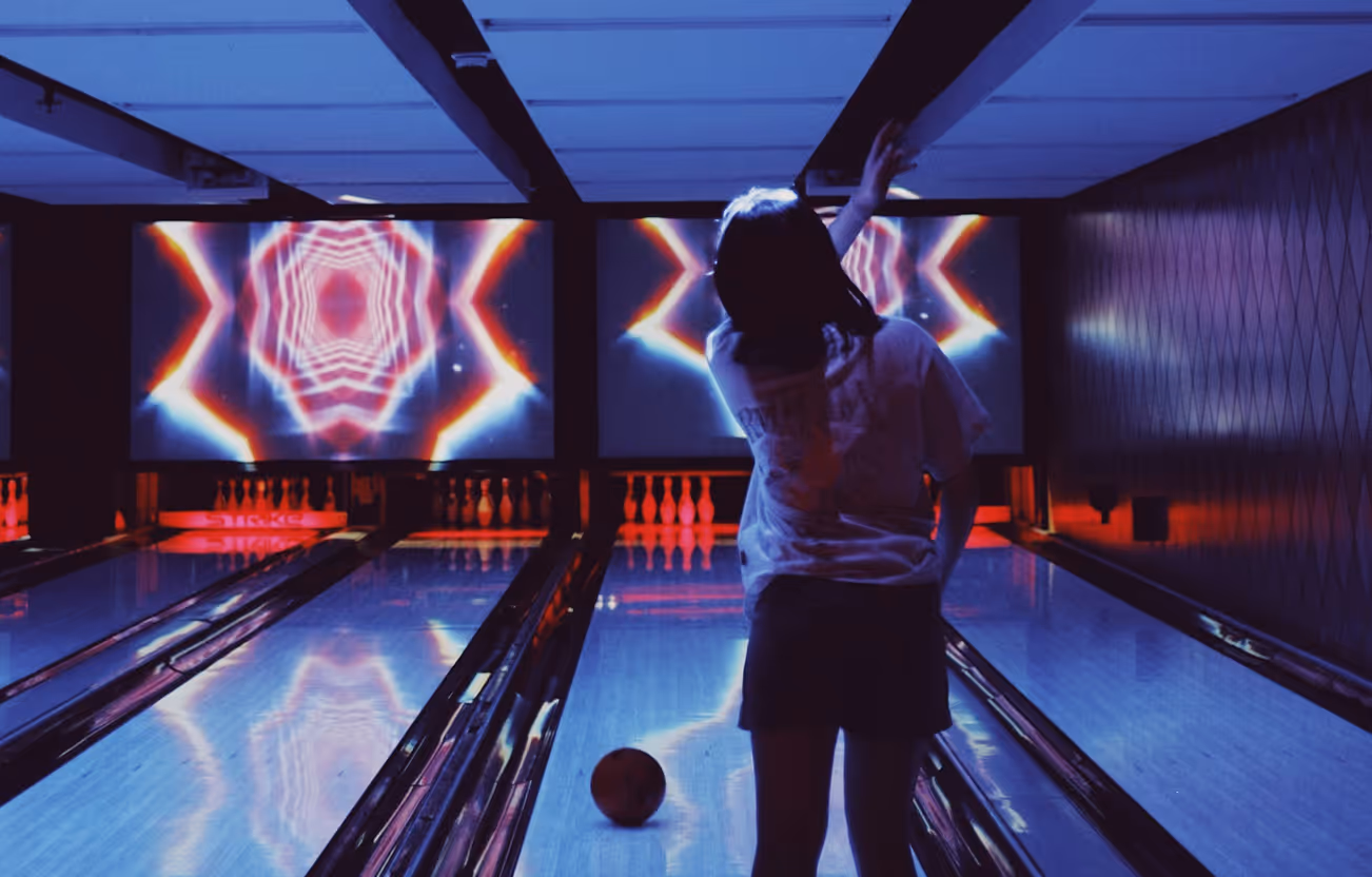 Person in casual clothes releasing a bowling ball down a lane in a dimly lit bowling alley with colorful psychedelic screen projections.