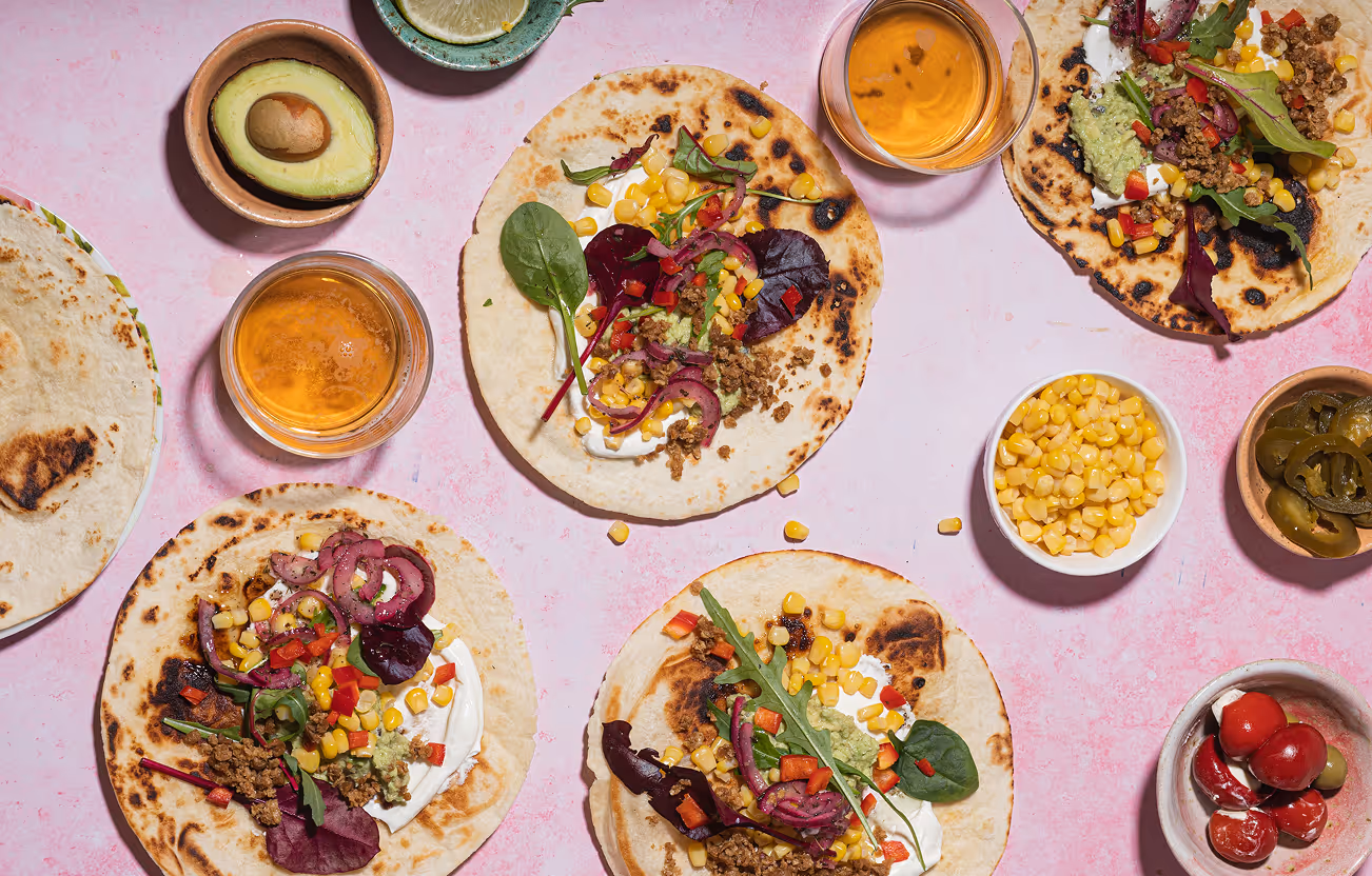 Four small tortillas topped with sour cream, cooked ground meat, corn, red bell peppers, pickled onions, guacamole, and mixed greens on a pink surface with bowls of corn, pickled jalapeños, avocado half, lemon slice, and cherry peppers.