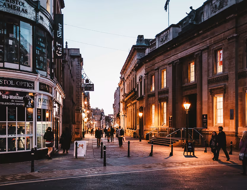 Evening street scene in Bristol with pedestrians walking along a historic building lit by street lamps.