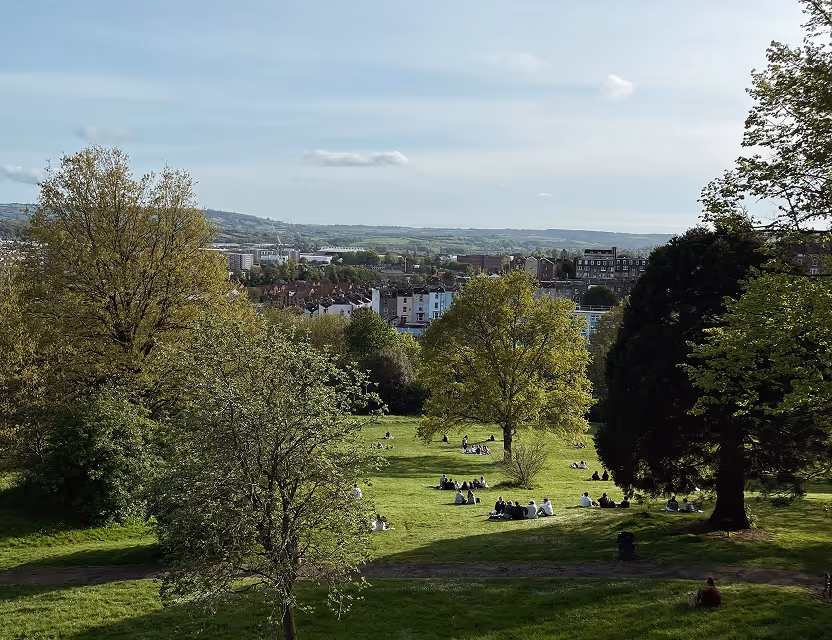 People sitting and relaxing on a large grassy area with scattered trees in a park overlooking a town and rolling hills under a partly cloudy sky.