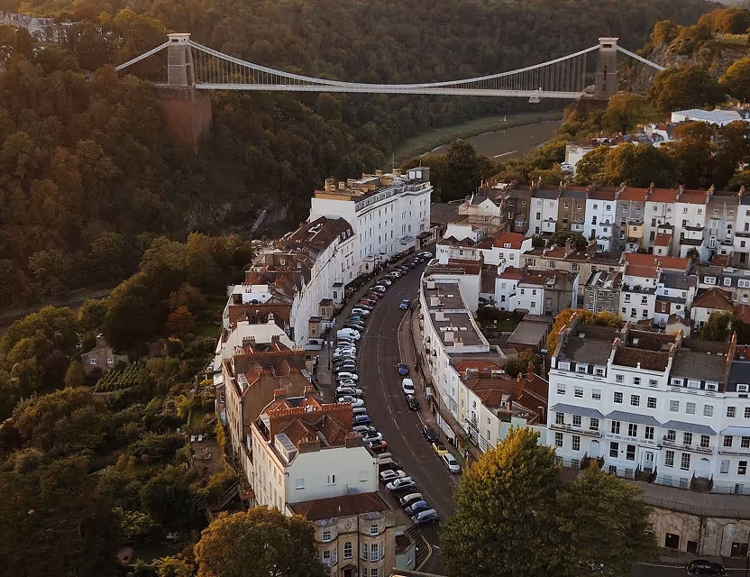 Aerial view of a curved street lined with parked cars and white buildings, with a suspension bridge spanning a green river valley in the background.
