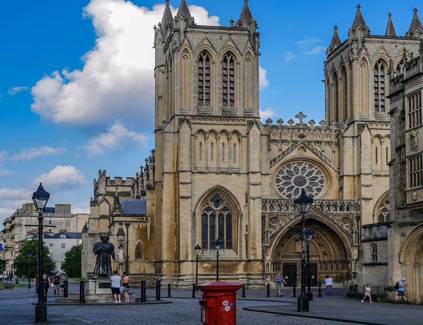 Gothic-style cathedral with large ornate windows and entrance, surrounded by people and street lamps on a sunny day.