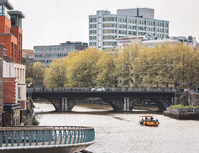Boat with people cruising on a river under a stone arch bridge with buildings and trees in the background.