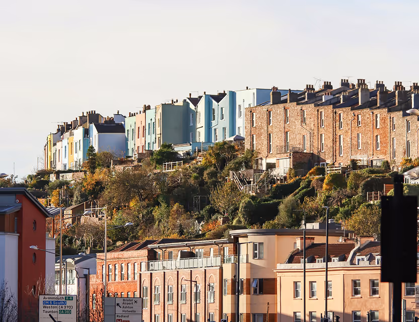 Colorful row houses on a green hillside overlooking beige buildings in an urban area under a clear sky.