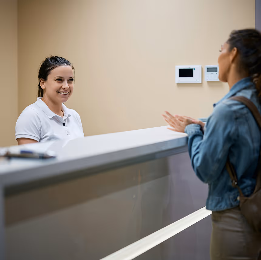 Smiling receptionist in white shirt assisting a visitor standing at the front desk in an office.