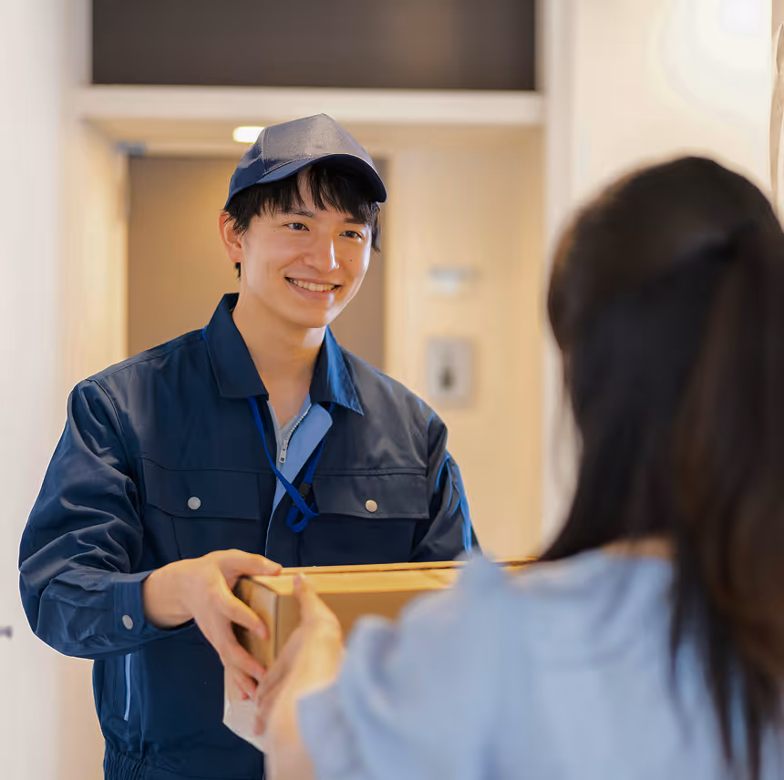 Smiling delivery man in navy uniform handing a cardboard package to a woman at her door.