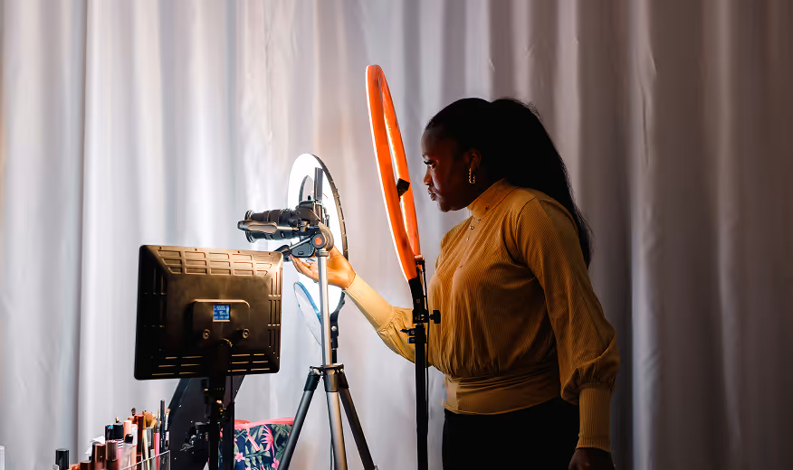 Woman adjusting a camera mounted on a tripod next to a ring light and monitor, in a well-lit indoor setup.
