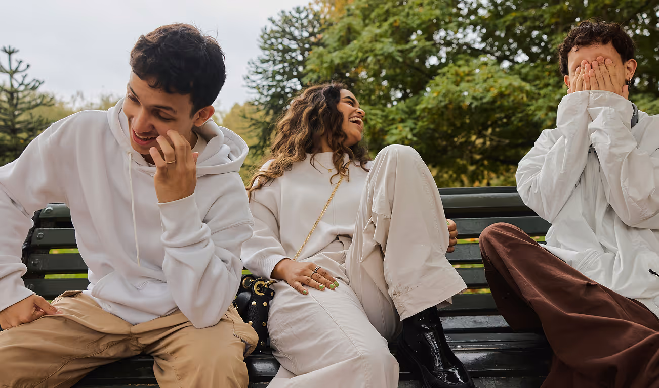 Three friends sitting on a park bench laughing and playing; one covers his face, another smiles looking down, and a woman in white laughs with her leg up.