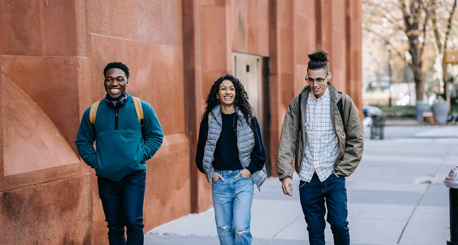 Three young adults smiling and walking on a city sidewalk beside a red brick building.