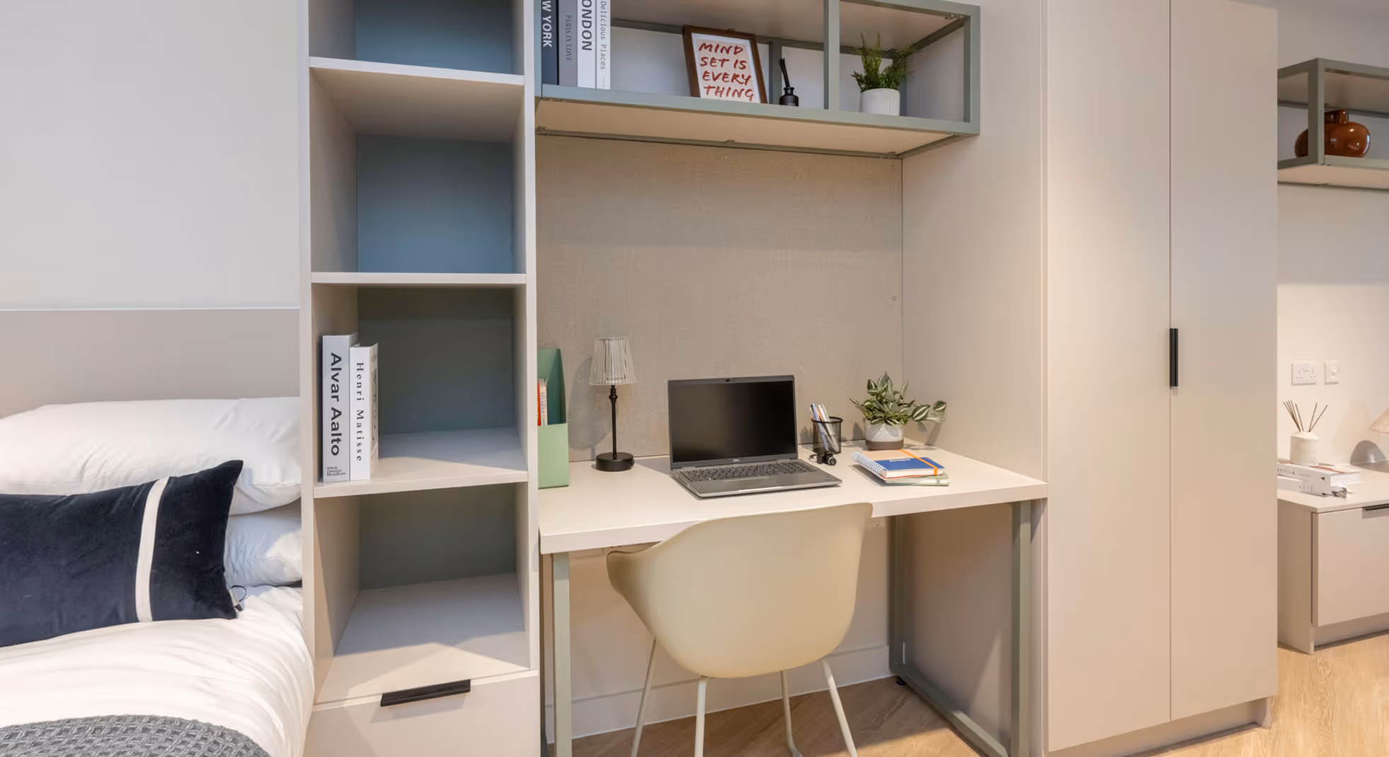 Modern study desk with laptop, lamp, books, and plant next to a bed with white and dark navy pillows in a tidy room.