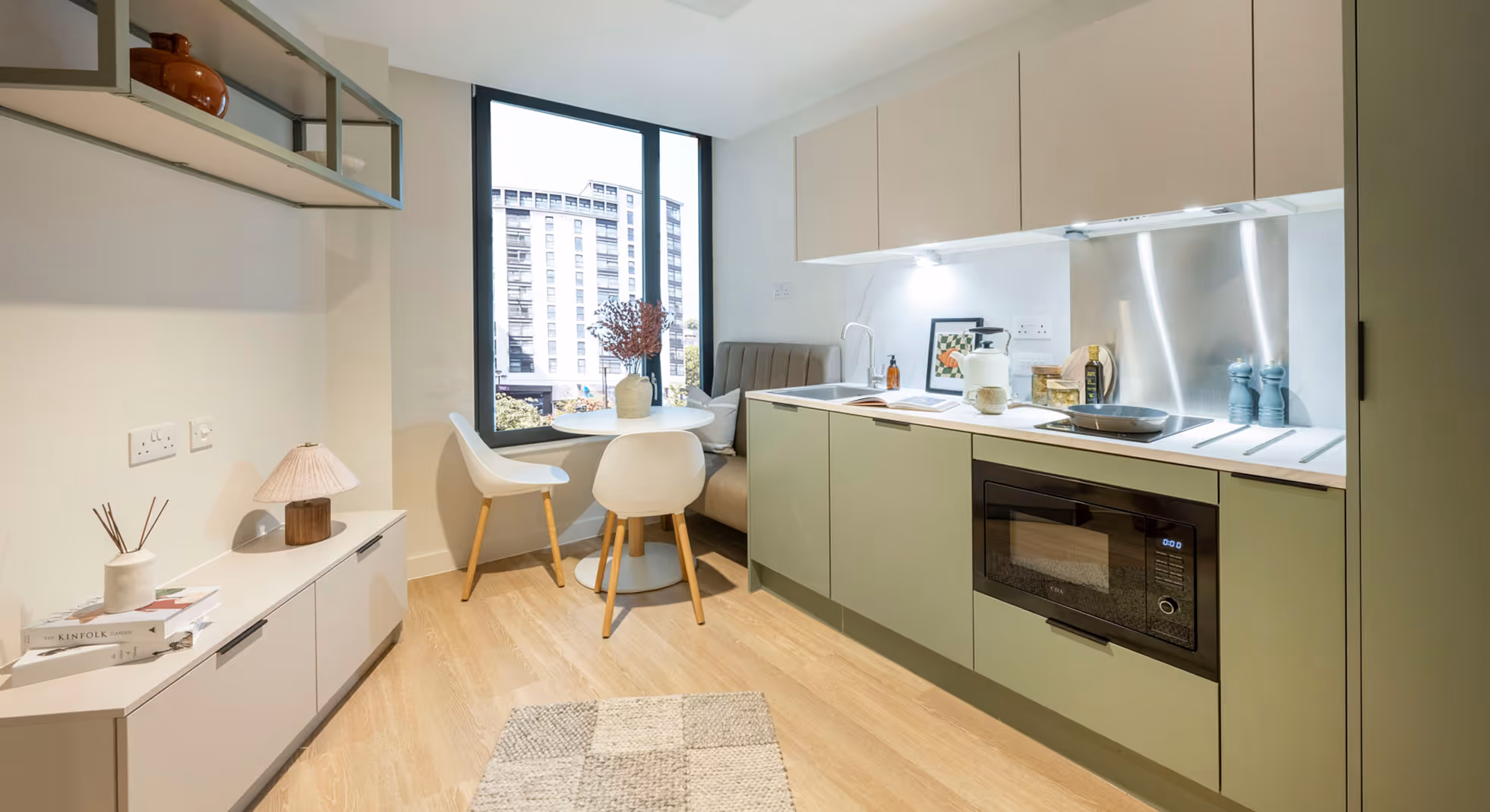 Modern kitchen with green cabinets, built-in microwave, small round dining table with white chairs, and large window showing a city building.