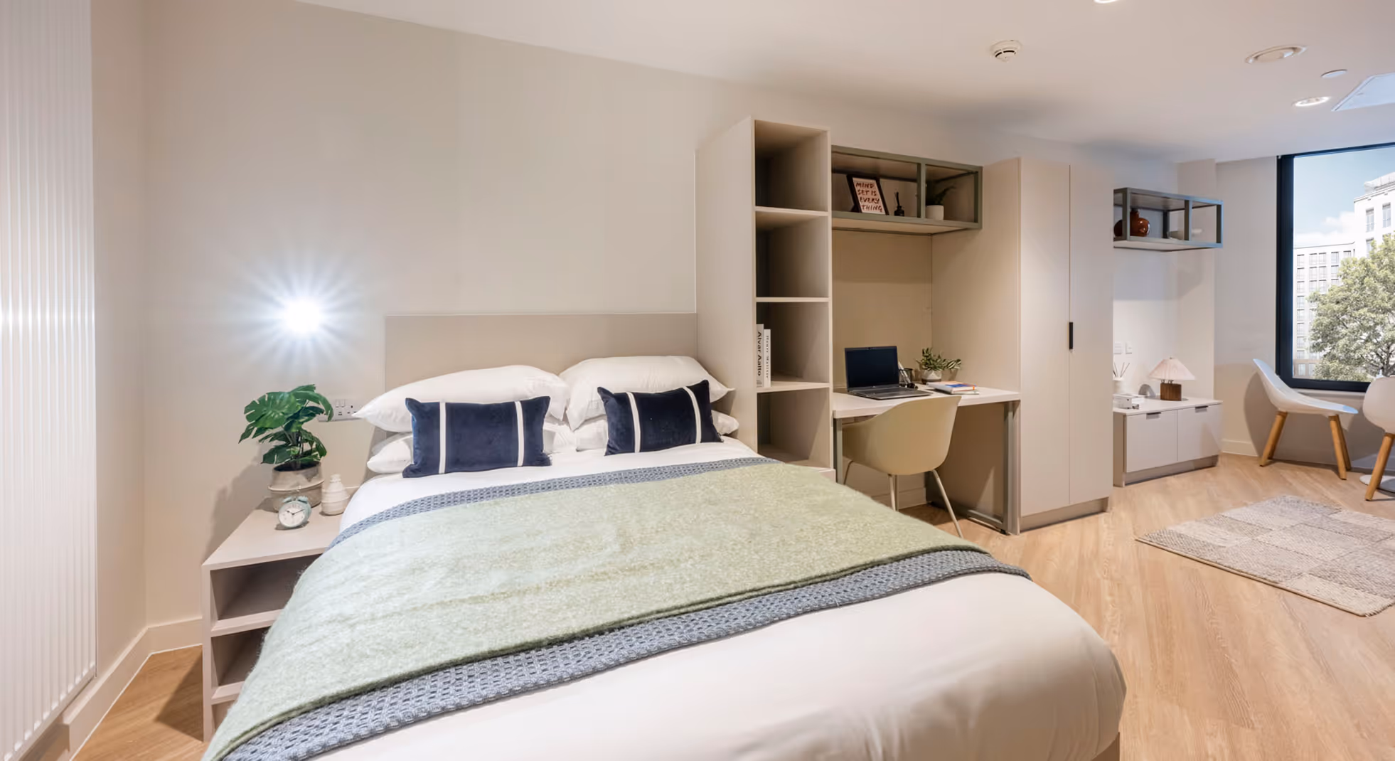 Modern bedroom with a neatly made bed, green throw blanket, two navy accent pillows, a desk with a laptop, and a large window showing city buildings and trees.