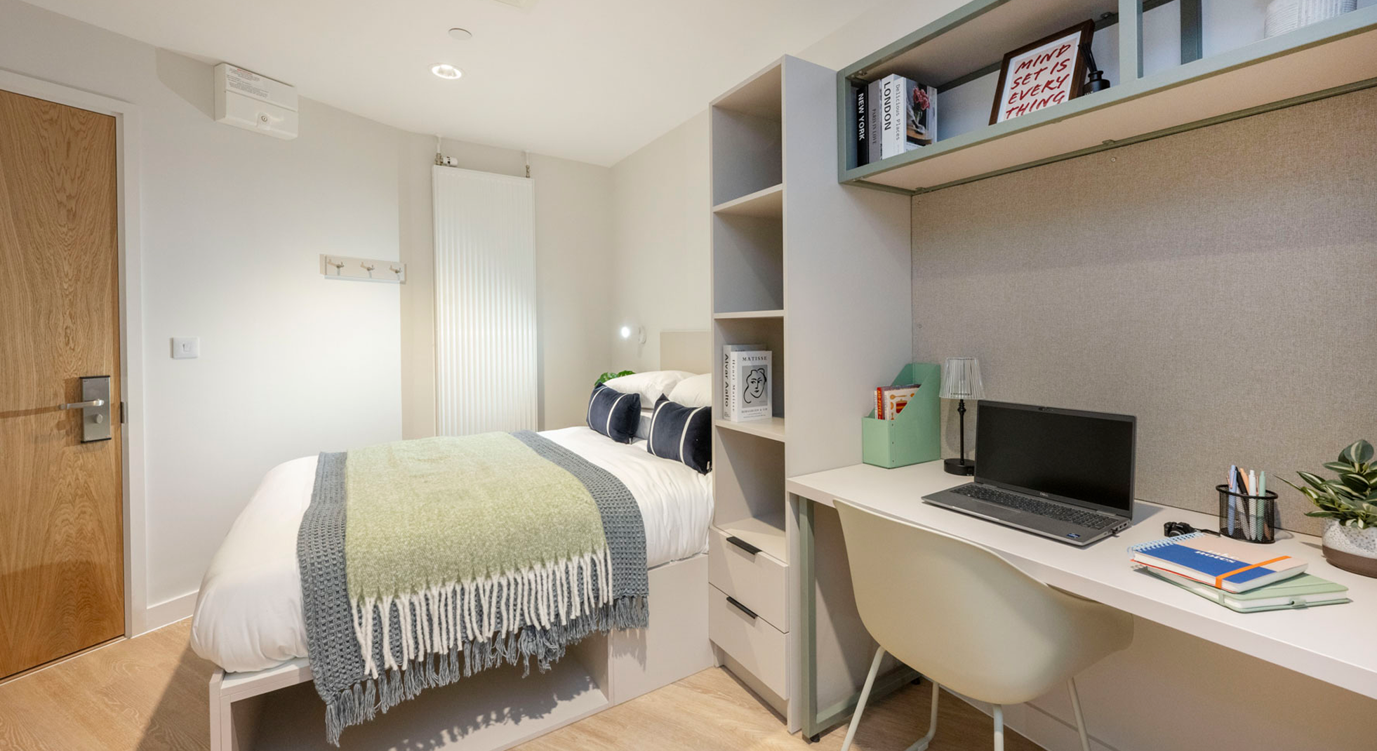 Minimalist bedroom with a double bed featuring white and blue pillows, a green throw, and an adjacent desk with a laptop, books, and a chair.