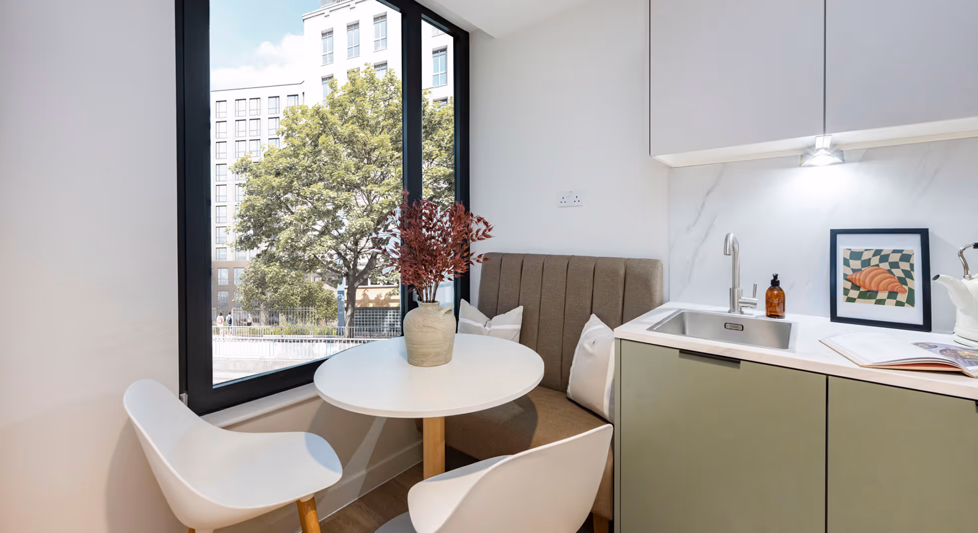 Bright modern kitchenette with round white table, two white chairs, beige cushioned bench, and a large window overlooking trees and buildings.